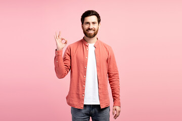 Happy young bearded man showing ok gesture looking at camera against pink background