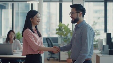 asian young woman and a asian man shaking hands in an office setting. The woman is wearing a pink blouse and black pants, and the man has a beard and glasses