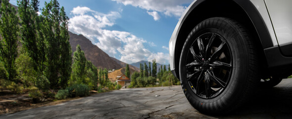 A black tire on a white car is shown close up on a rocky road with mountains and river in the background.