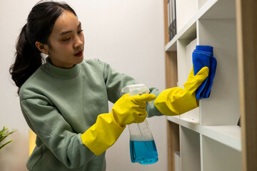 A woman is cleaning a bookshelf with a yellow brush. She is wearing rubber gloves and a green sweater
