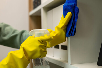 A woman is cleaning a bookshelf with a yellow brush. She is wearing rubber gloves and a green sweater
