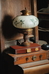 Vintage still life featuring ornate table lamp with white glass shade and floral pattern, sitting atop antique wooden boxes with brass hardware and worn patina. Generative AI