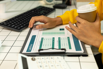 A woman in a yellow sweater is sitting at a desk with a computer monitor and a clipboard. She is...