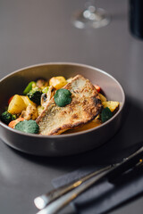 Crispy seabass fillet with roasted vegetables, creamy bisque sauce, and fresh herbs, elegantly plated in a dark ceramic bowl on a stylish minimalistic table setting