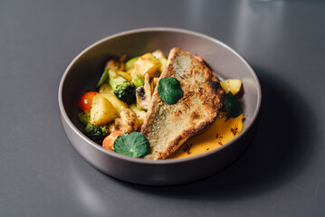 Gourmet dish with crispy seabass fillet, roasted vegetables, and saffron-infused bisque sauce, elegantly plated in a ceramic bowl on a dark table setting