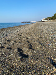 Scenic pebble beach with footprints, calm sea, distant pier, and a clear blue sky on a sunny day.