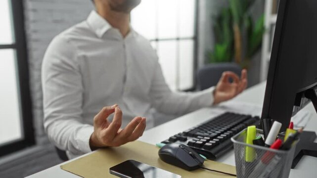 Man meditating at office desk with computer, taking deep breath during work break, practicing mindfulness for stress relief indoors, illustrating modern workspace environment.