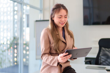 Fototapeta premium Young Asian businesswoman smiling while using a tablet in a bright and modern office environment