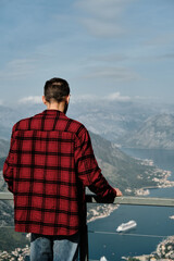 Fototapeta premium A young Caucasian man in a red plaid shirt stands on an observation deck overlooking the Bay of Kotor. Rear view. The southernmost fjord in the Adriatic Sea, Montenegro. Travel concept