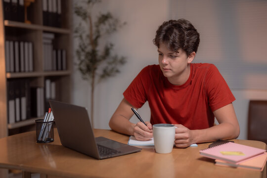Young man attending an online class, sipping coffee and writing notes in a notebook while seated at a desk with a laptop, surrounded by books on a bookcase