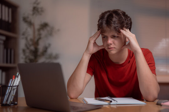 Overworked teenager having headache and massaging his temples while using laptop for studying online at home, concept of stress, exhaustion, overwork, education and learning difficulties