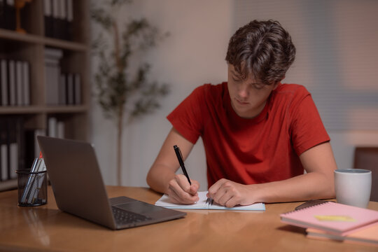 Concentrated teenager writing notes in a notebook while studying at home during the night, surrounded by a laptop and a steaming cup of coffee on the desk