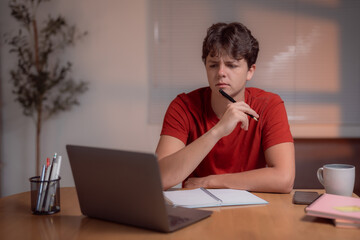 Young student attending online class using laptop and taking notes on notebook, sitting at home at desk with laptop, notebook, pen, smartphone and coffee mug