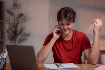 Happy male student using mobile phone and taking notes while studying with laptop at home, managing his time efficiently and multitasking between communication and learning