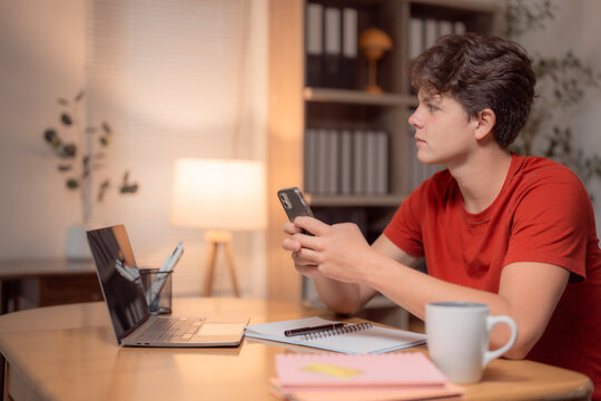 Young man studying at a desk, using a mobile phone while working on a laptop surrounded by notebooks. Nighttime ambiance fills the cozy room, enhancing focus and concentration - Powered by Adobe