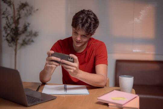 Young man in a red shirt focusing on a smartphone while studying at a desk. A laptop, notebooks, and a mug are present, suggesting a multitasking environment in a home office