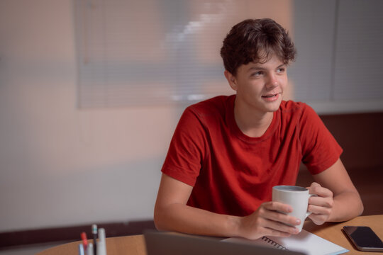 Young man in a casual red shirt holds a coffee mug, taking a break at a desk with stationery and a laptop. He appears thoughtful and relaxed, enjoying a moment of leisure