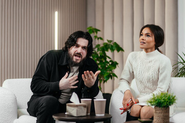 Multiracial couple man and woman sitting on the couch and arguing with each other. Quarrel at a psychologist's appointment. Angry man, waving his arms aggressively. Abuse in relationships