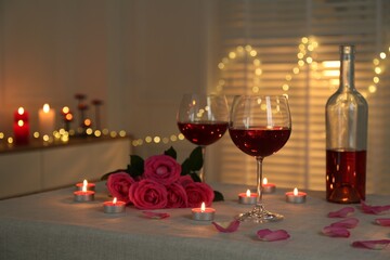 Romantic place setting with red wine, burning candles and roses on table against blurred lights