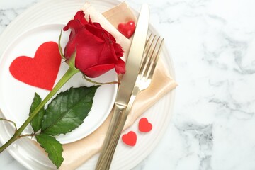 Romantic place setting for Valentine's day. Tableware, cutlery, rose and decorative hearts on white marble table, top view. Space for text
