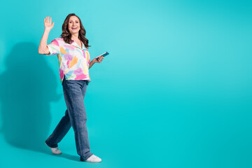 Full size photo of pretty retired female hold gadget walking wave hand wear trendy colorful outfit isolated on cyan color background