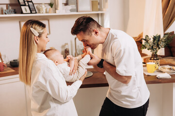 Fototapeta premium Happy family with newborn son playing in the kitchen. Family couple at home with son making coffee in the kitchen. Home family atmosphere