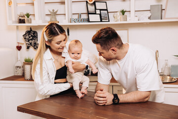 Smiling young family with child, father and mother with little baby, newborn child, having morning breakfast in home kitchen. Family enjoying parenthood at home.