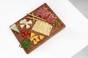 Cheese wooden board on white table. Different types of cheese, chili, jamon, sausage, olive, tomato, microgreens, knife, breadsticks. Snack. Pieces of cheese.