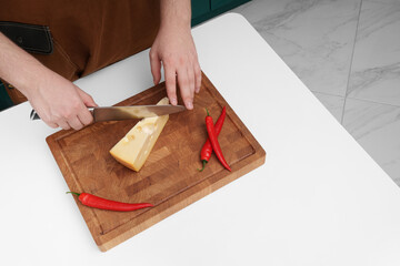 A man in the kitchen cuts cheese on a board on a white table. Cheese, chili, knife. Appetizers. Slicing.
