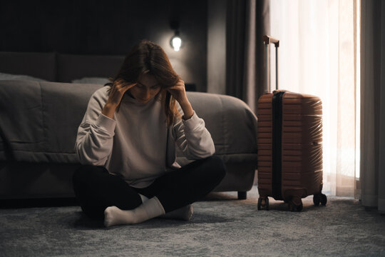 Depressed lonely woman sitting in hotel room with her suitcase nearby. Female emotional crisis during solo travel, feeling sense of loneliness, escape from complicated situation, or violence 