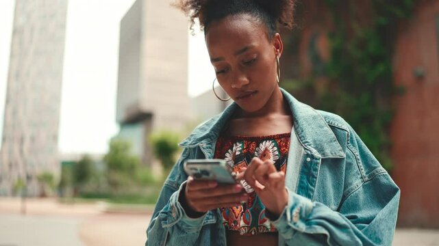 Close-up African girl wearing denim jacket, in crop top with national pattern goes on the street of city and holding smartphone looking at phone screen look for address with electronic map in city