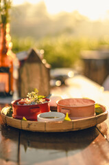 Close-up of breakfast container and bowl in a winnowing basket on a wooden table in the morning. Blurred background of mountain views and farms with copy space. Soft light shines down in warm tones.