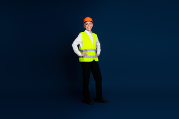 Confident mature woman in construction attire with orange hardhat and safety vest, stands against a dark blue background