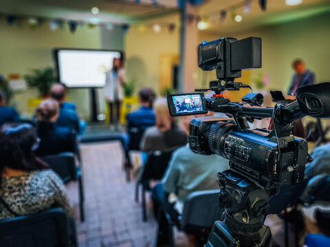 A professional video camera set up on a tripod captures a conference or seminar, with a blurred audience and speaker in the background. The camera screen shows