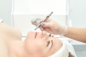 Caucasian female receiving facial treatment with airbrush device in spa setting.