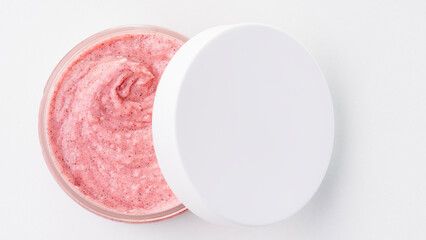 Pink body scrub in an open jar from above On a white background. The white lid is ajar and lies on the jar on top.