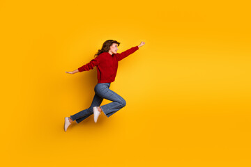 Fototapeta premium Young woman jumping gracefully against yellow background, wearing red sweater and denim jeans