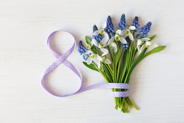 Wooden white background with a bouquet of spring flowers snowdrops and muscari, ribbon in the shape of the number eight. Postcard for Women's Day March 8th.