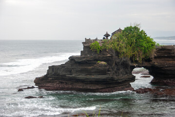 A scenic view of a rocky cliff with lush greenery, a traditional Balinese temple perched on top, and waves crashing against the shore, creating a tranquil atmosphere at the coastal landscape.