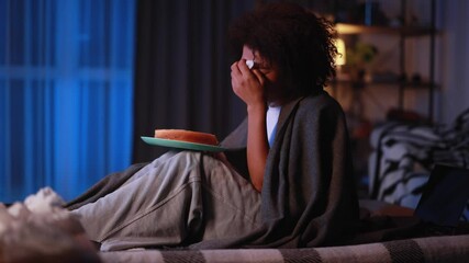 Sad young african american woman dealing with stress by eating food sitting on bed late at home Upset female wrapped in blanket crying and eating cake at night indoors alone Mental heath problem