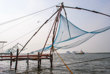 A traditional Chinese fishing net set in the water, stretching across the surface. A bird flies above while a cargo ship can be seen in the background, adding contrast to the rural fishing method.