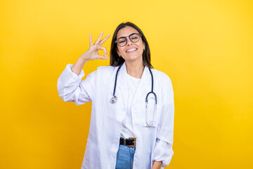 Young brunette doctor woman wearing stethoscope standing over isolated yellow background doing ok...