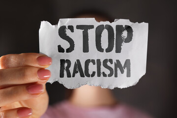 Woman holding piece of paper with text Stop Racism on dark background, closeup