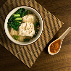 Wonton dumpling soup, A delicious bowl of dumpling soup featuring fresh greens, served on a bamboo mat with a side of spicy sauce for added flavor