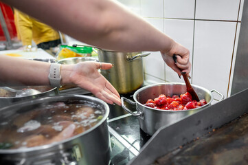 A hand stirs freshly prepared strawberries in a pot on a kitchen stove, surrounded by other cooking pots. Warm, inviting atmosphere highlights culinary activity.