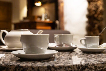 Elegant tea and coffee setup with white porcelain cups and a teapot on a granite table in a cozy cafe setting with warm ambient lighting. Cozy coffeeshop scene. Selective focus
