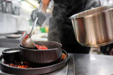A chef serves a steaming bowl of flavorful soup in a modern kitchen. The dish is garnished with spices, highlighting culinary artistry and the vibrant atmosphere of food preparation.