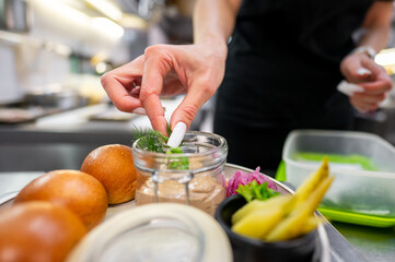A chef's hand garnishes a dish with fresh herbs. The plate features small rolls, pickles, and a creamy dip, showcasing a focus on culinary presentation and attention to detail.