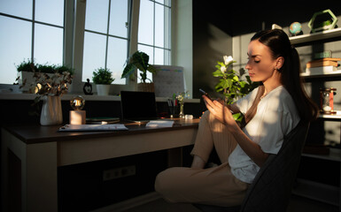 A woman in a white t-shirt and beige pants sits at a desk, looking at her phone. The warm sunlight...