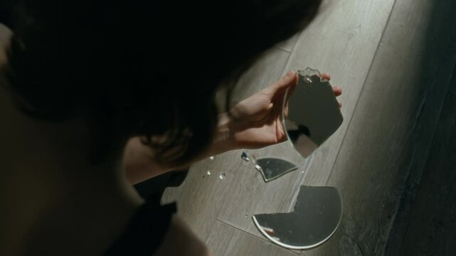 Over the shoulder selective focus shot of stressed girl looking at her reflection in shard of broken mirror on floor